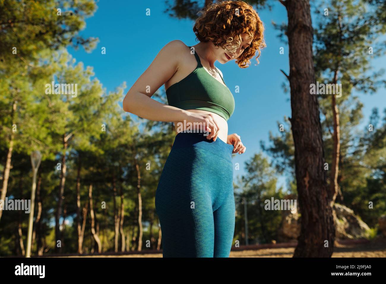 Cute redhead woman wearing green sports bra and blue yoga pants standing on  city park, outdoors she holds hands at the waist. Flat stomach, healthy li  Stock Photo - Alamy
