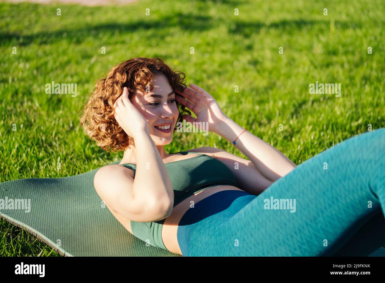 Young happy redhead woman wearing sportive clothes doing sit-ups abs crunches exercise indoors lying on yoga mat. Sport workout routine, morning physi Stock Photo