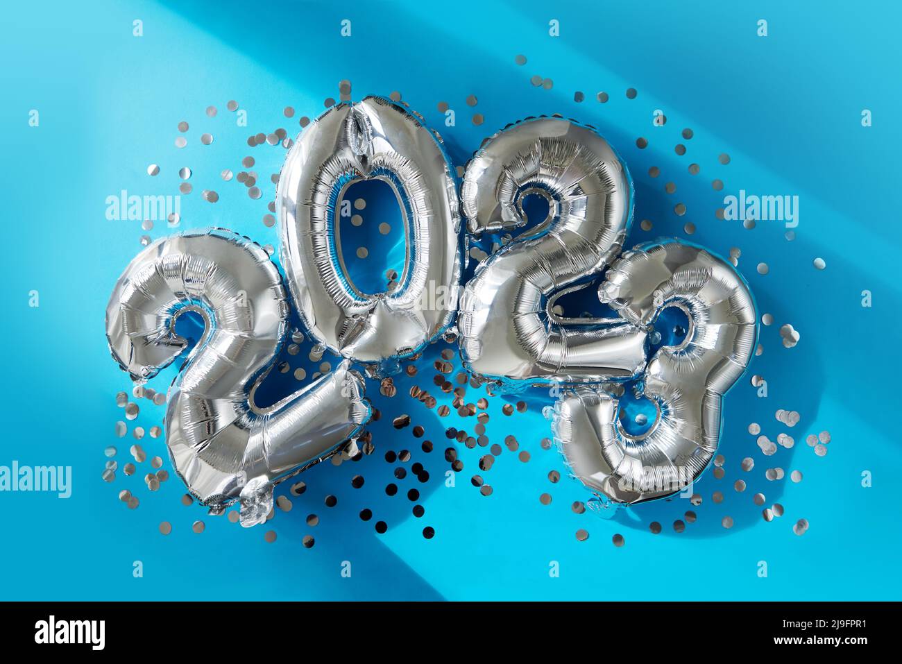 Silver numbers 2023 new year balloons among confetti in sunlight on blue background Happy new year celebration party. Greetings and congratulation Stock Photo