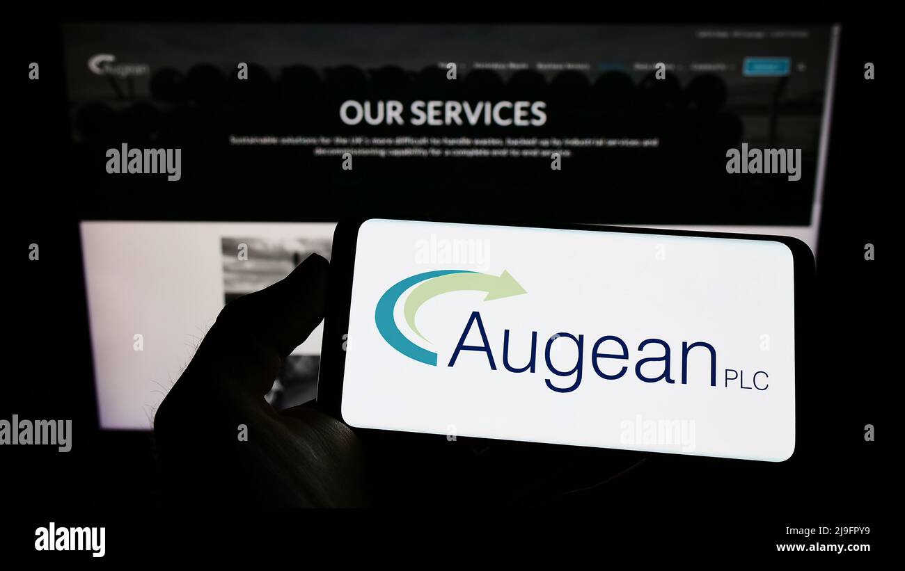 Person holding mobile phone with logo of British waste management company Augean plc on screen in front of web page. Focus on phone display. Stock Photo