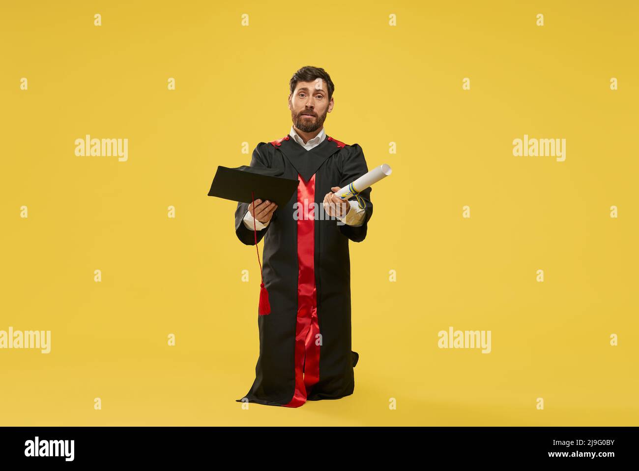 Front view of student with beard standing on knees, begging. Master, bachelor wearing graduate gown, holding diploma and mortarboard. Isolated on yellow studio background. Stock Photo
