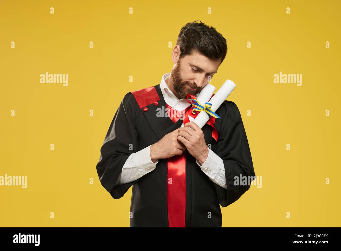 Front view of student holding, hugging two diplomas. Brunette boy with beard, wearing graduate gown, graduating from college, looking down. Isolated on yellow studio background. Stock Photo