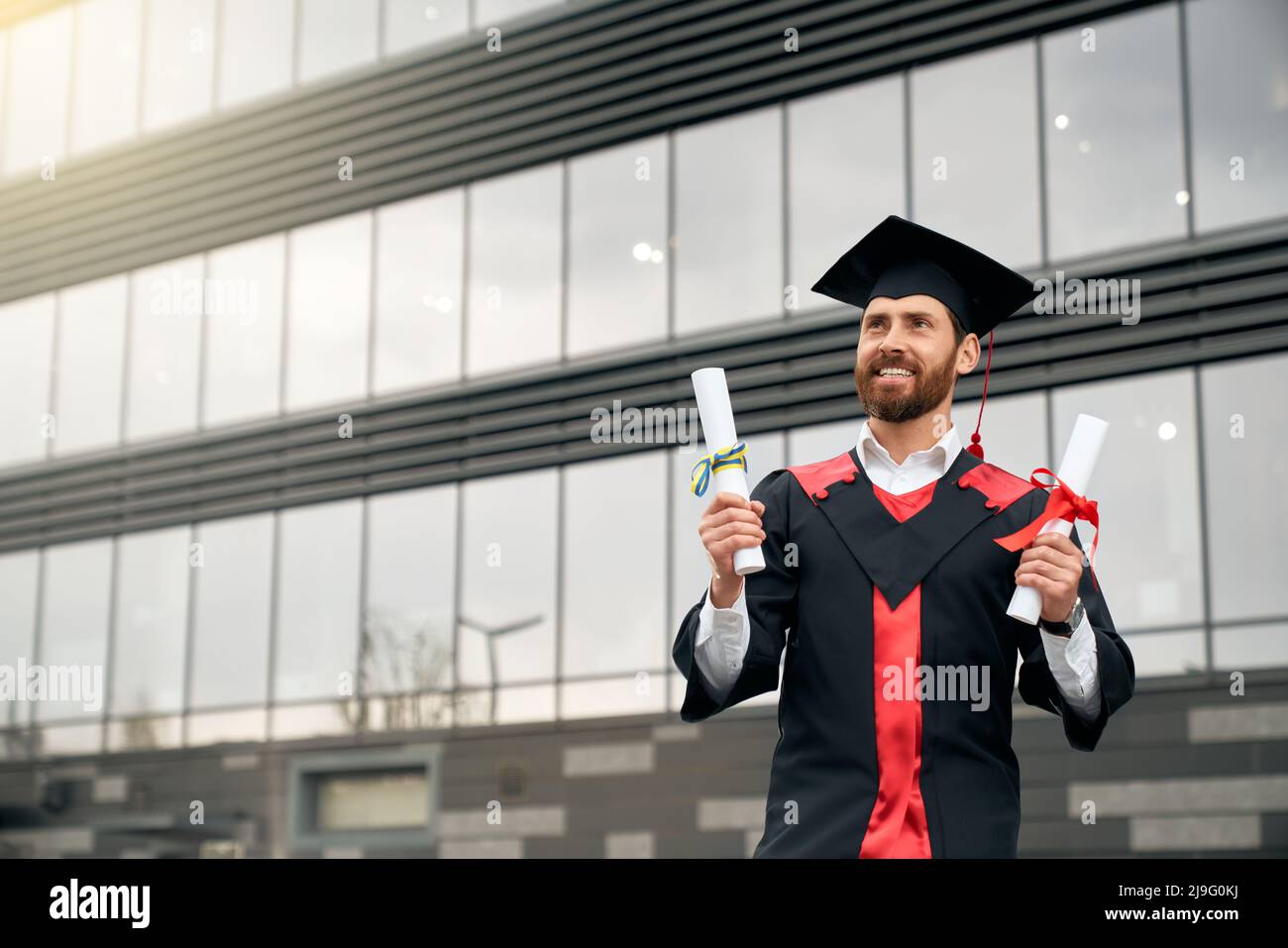 Front view of student in mortarboard and graduate gown holding two diplomas. Handsome young man graduating from college, smiling, happy, cheerful. Concept of education and youth. Stock Photo