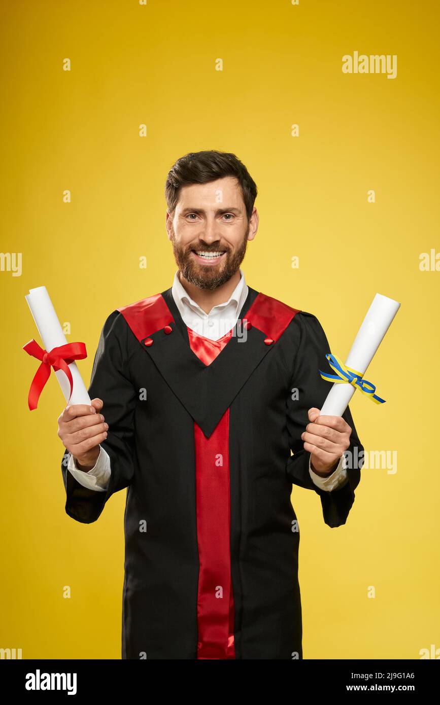 Front view of student with beard standing, holding two diplomas. Master, bachelor graduating from university, college, smiling, looking at camera. Isolated on yellow background. Stock Photo