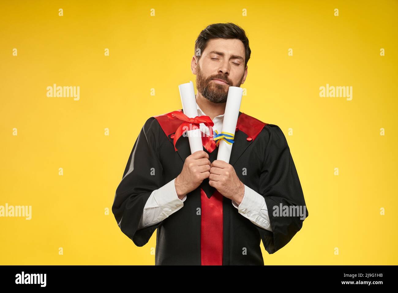 Front view of bachelor, master standing, holding two diplomas. Student wearing graduate gown and mortarboard, happy, glad with closed eyes. Isolated on yellow studio background. Stock Photo