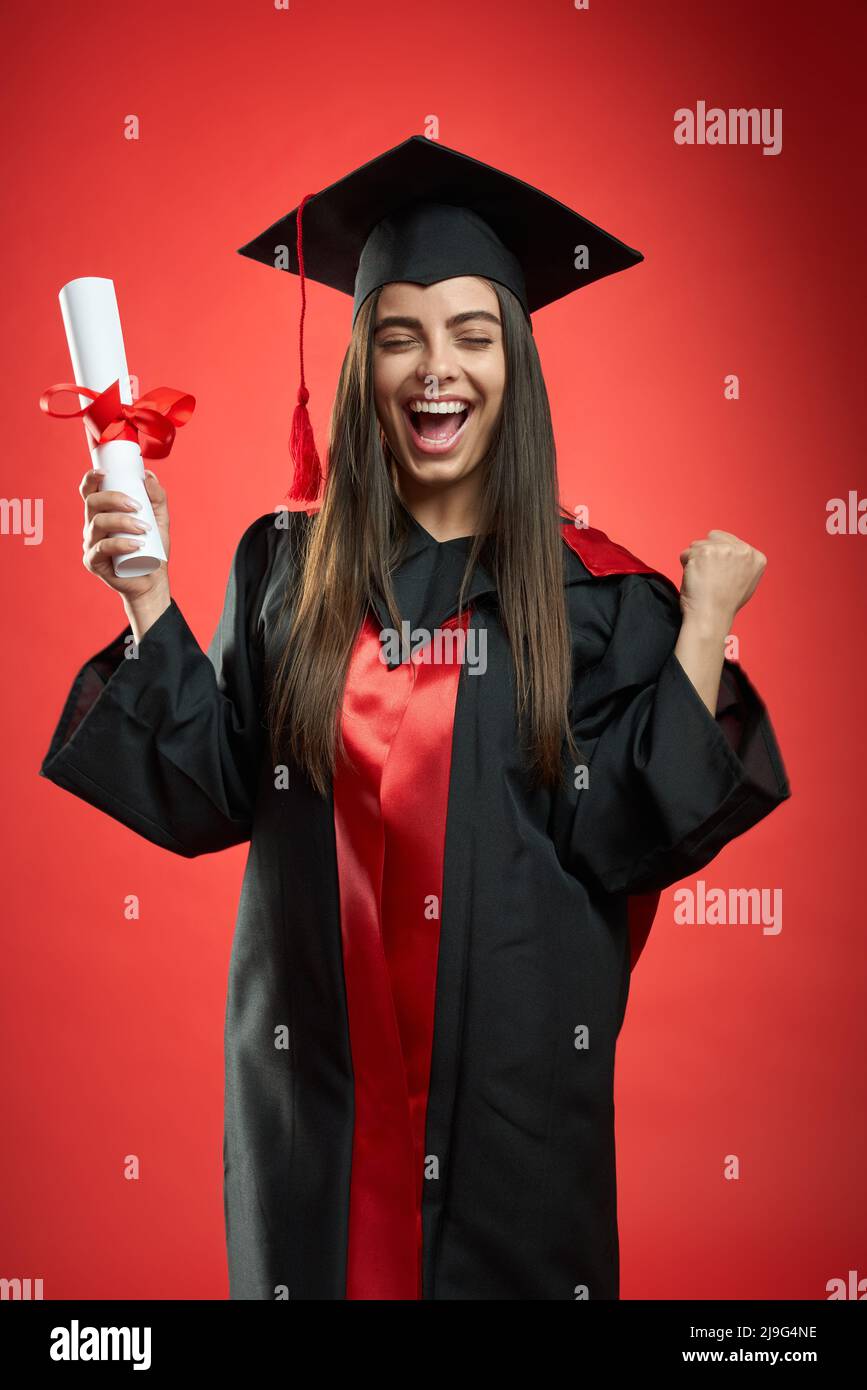 Front view of glad, happy girl graduating from college, university, high school. Pretty young female standing with closed eyes, holding diploma, laughing. Concept of education. Stock Photo