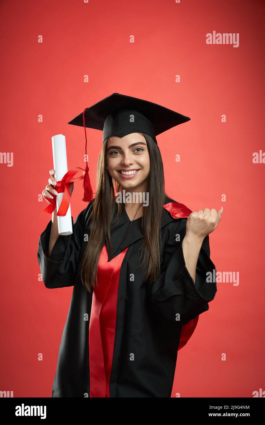 Front view of pretty girl in graduate gown and mortarboard graduaing from college. Young female standing, holding diploma, showing hurray, smiling. Concept of education and youth. Stock Photo