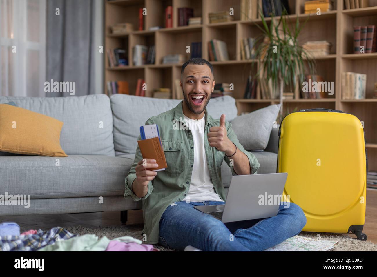 Online hotel reservation concept. Happy black man with passport and tickets using laptop, showing thumb up gesture Stock Photo