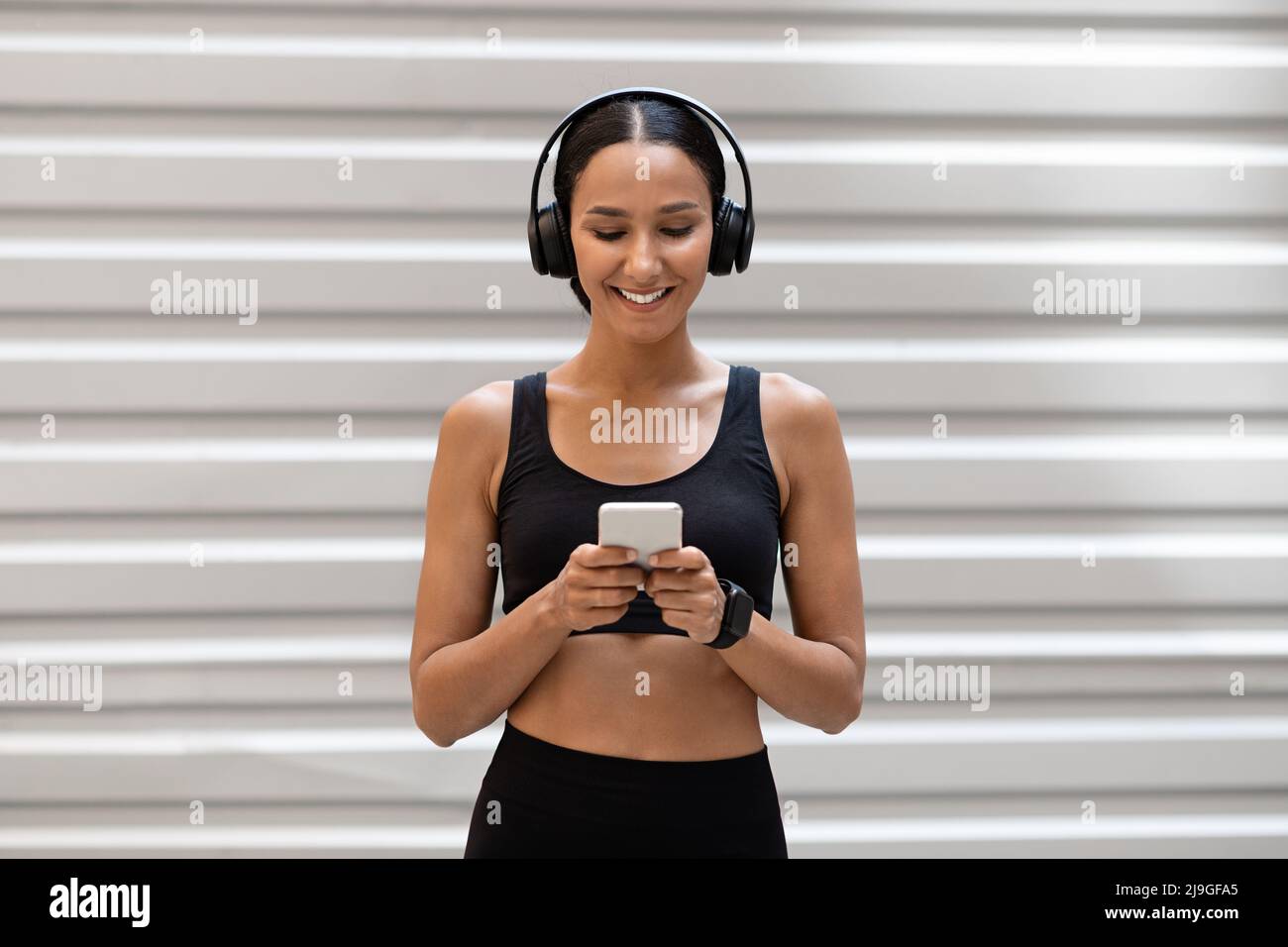 Cheerful young slim arab female in sports uniform and headphones typing on smartphone on gray wall background Stock Photo