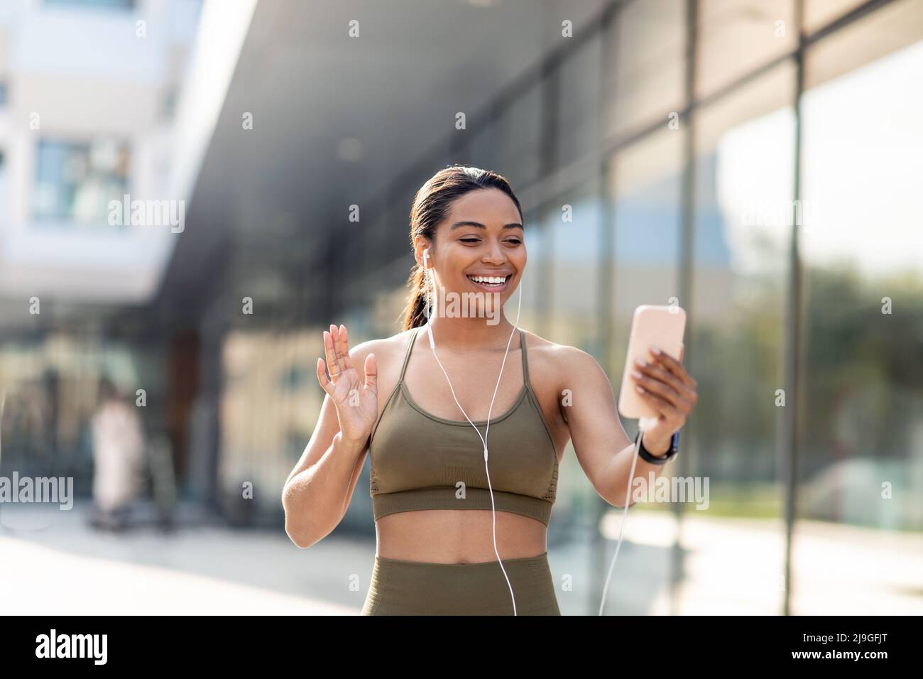 Athletic young black lady waving at smartphone screen, having online video call with fitness trainer on city street Stock Photo