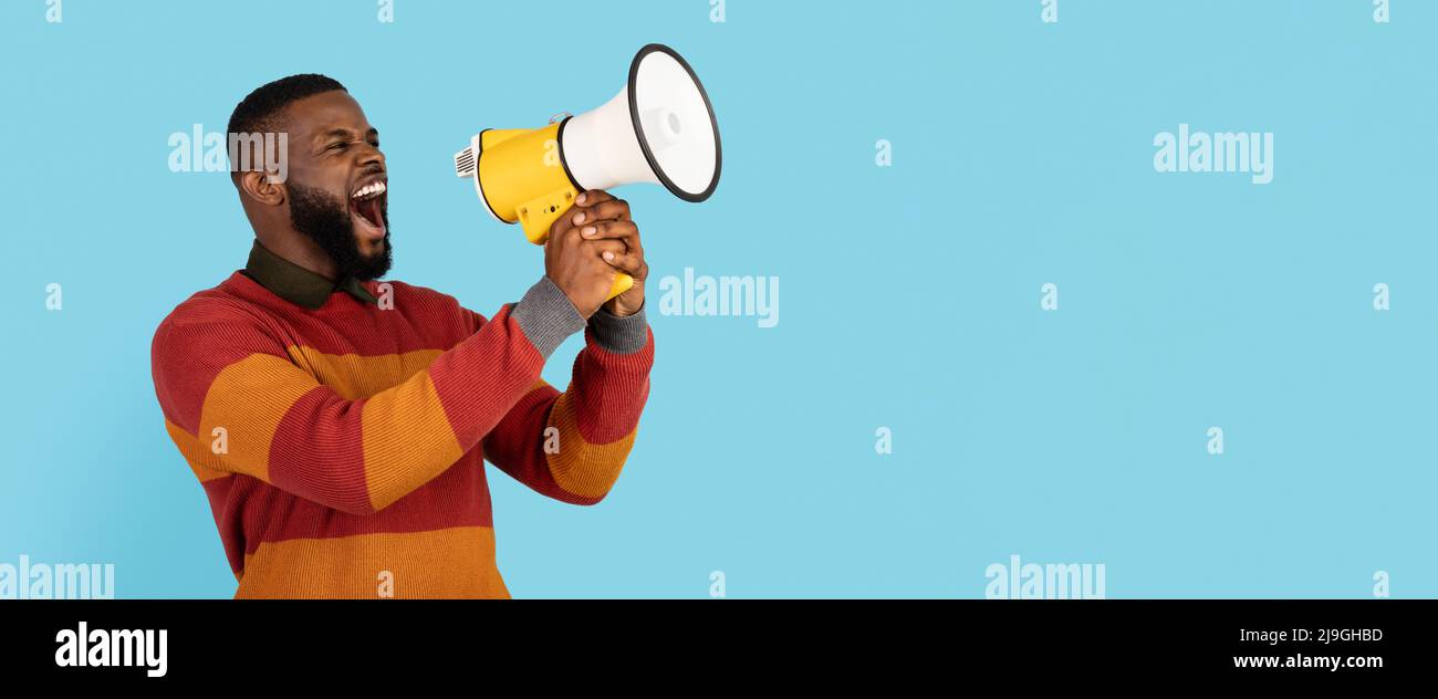 Portrait Of Young African American Man With Megaphone In Hands Making Announcement, Black Millennial Male Shouting Aside At Copy Space On Blue Stock Photo