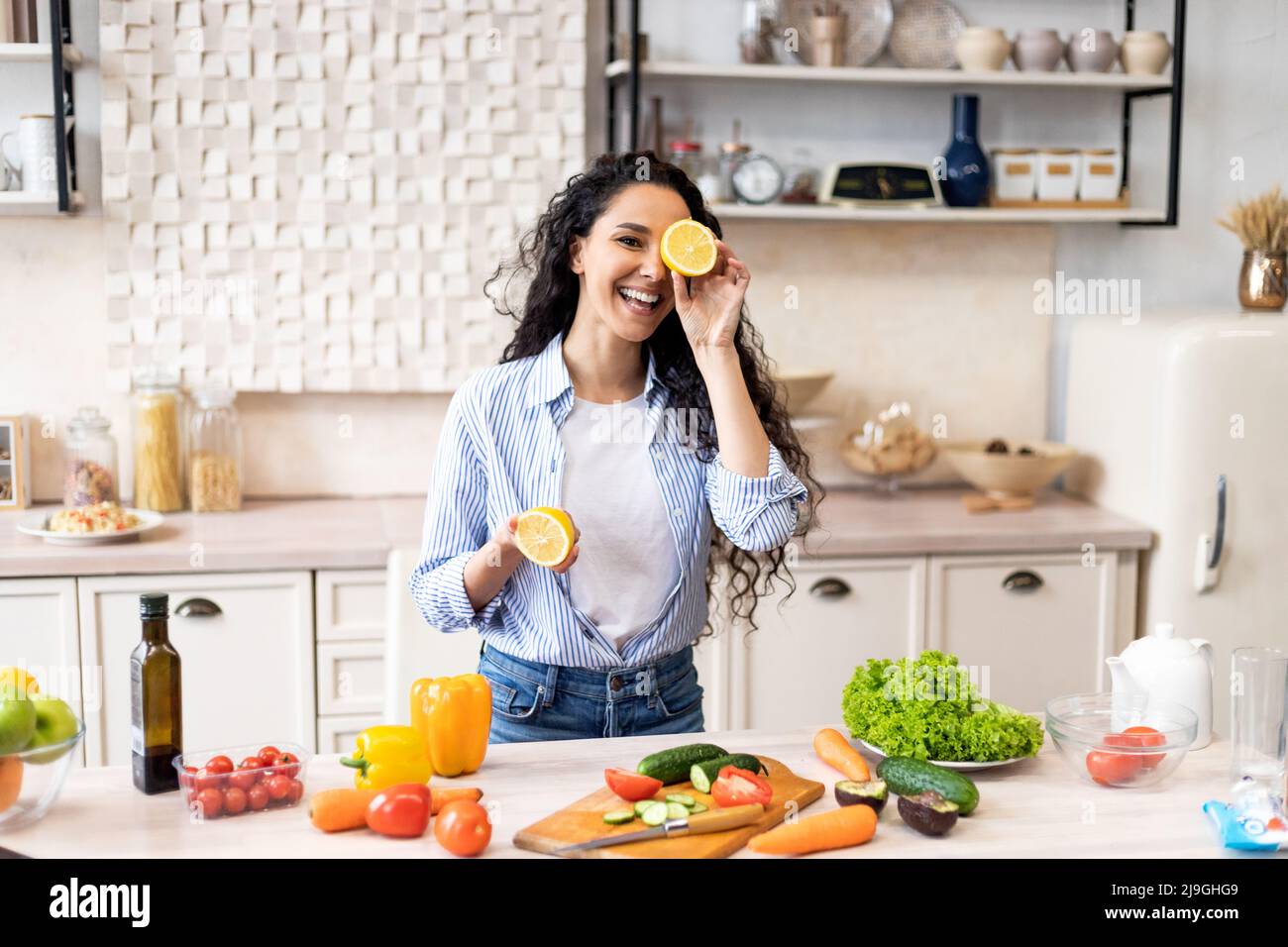 Happy young latin lady having fun and covering eyes with lemon halves, fooling around in kitchen while cooking dinner Stock Photo