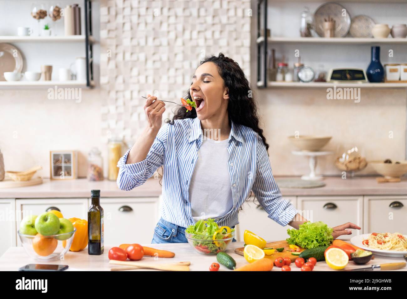 Young woman eating salad at table with organic vegetables, enjoying healthy diet, standing in light kitchen interior Stock Photo