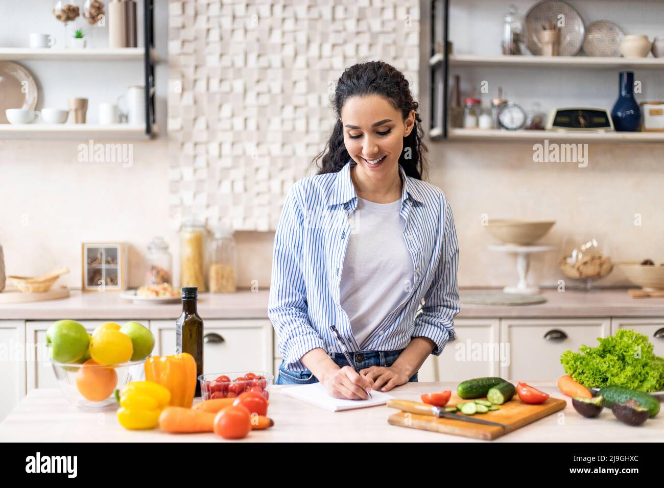 Happy young lady making notes on table with organic vegetables, cooking dinner in light kitchen interior Stock Photo