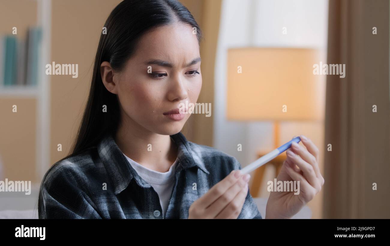 Puzzled doubtful Asian woman waiting result of pregnancy test girl worry with unplanned children. Sad unhappy pregnant Korean lady sitting at home Stock Photo