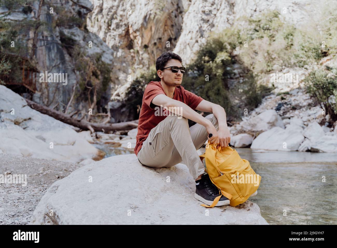 Man with yellow backpack sits on a riverside rock in the canyon with mountain cliffs in the background. Adult man taking a rest on a boulder near Stock Photo