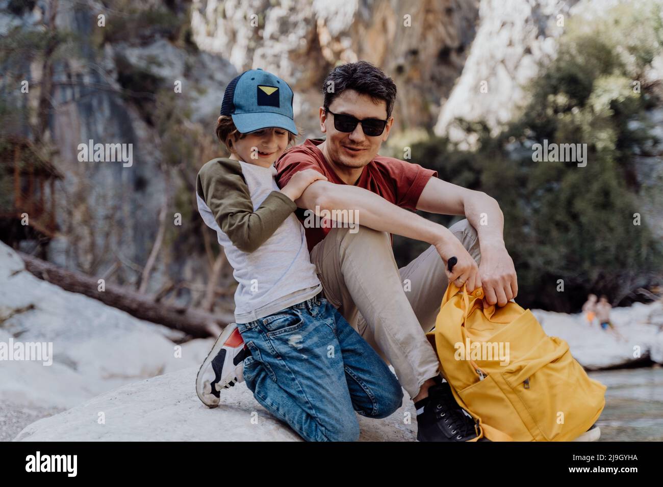 School boy and his dad with yellow backpack sits on a riverside rock in the canyon with mountain cliffs in the background. Kid child and father taking Stock Photo