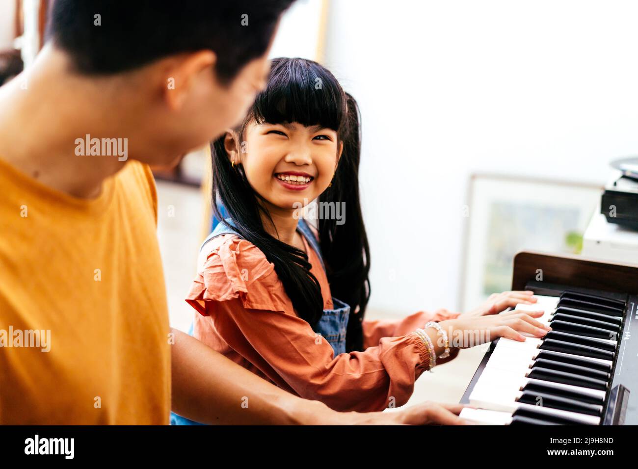 Cheerful Asian daughter in casual clothes with pigtails smiling and looking at dad while leaning to play piano in daytime at home Stock Photo