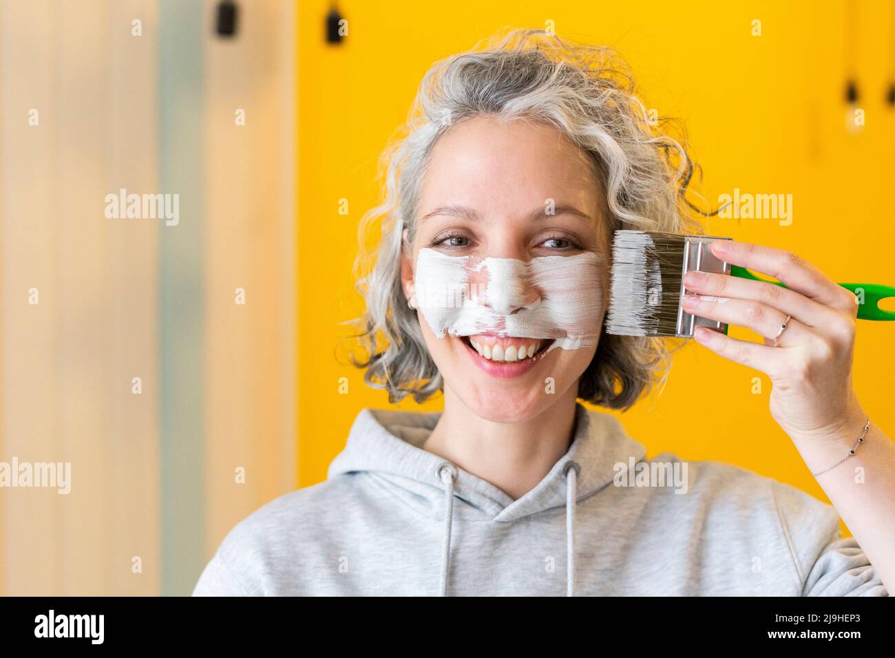 Smiling playful woman applying white paint on face with paintbrush Stock Photo