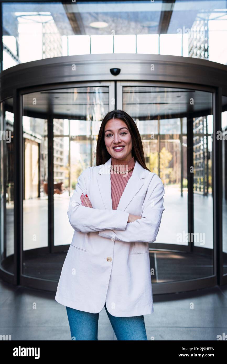 Happy businesswoman with arms crossed standing in front of revolving door Stock Photo