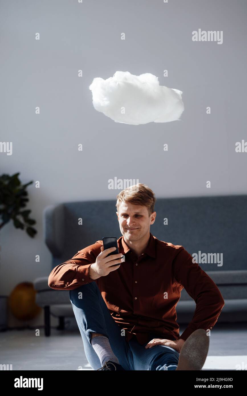 Smiling businessman using smart phone sitting under levitating cloud network in office Stock Photo