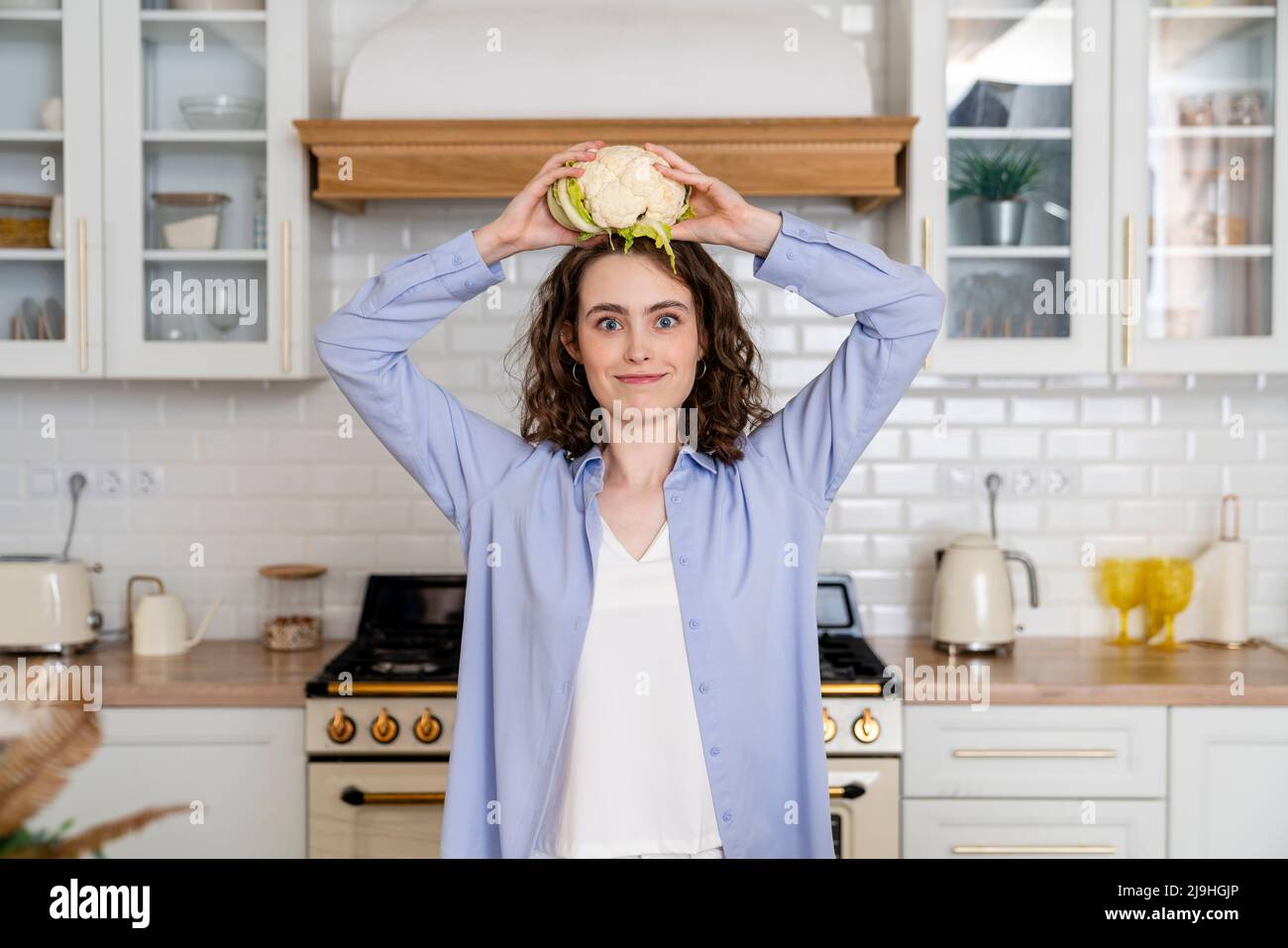 Young woman holding cauliflower on head in kitchen at home Stock Photo