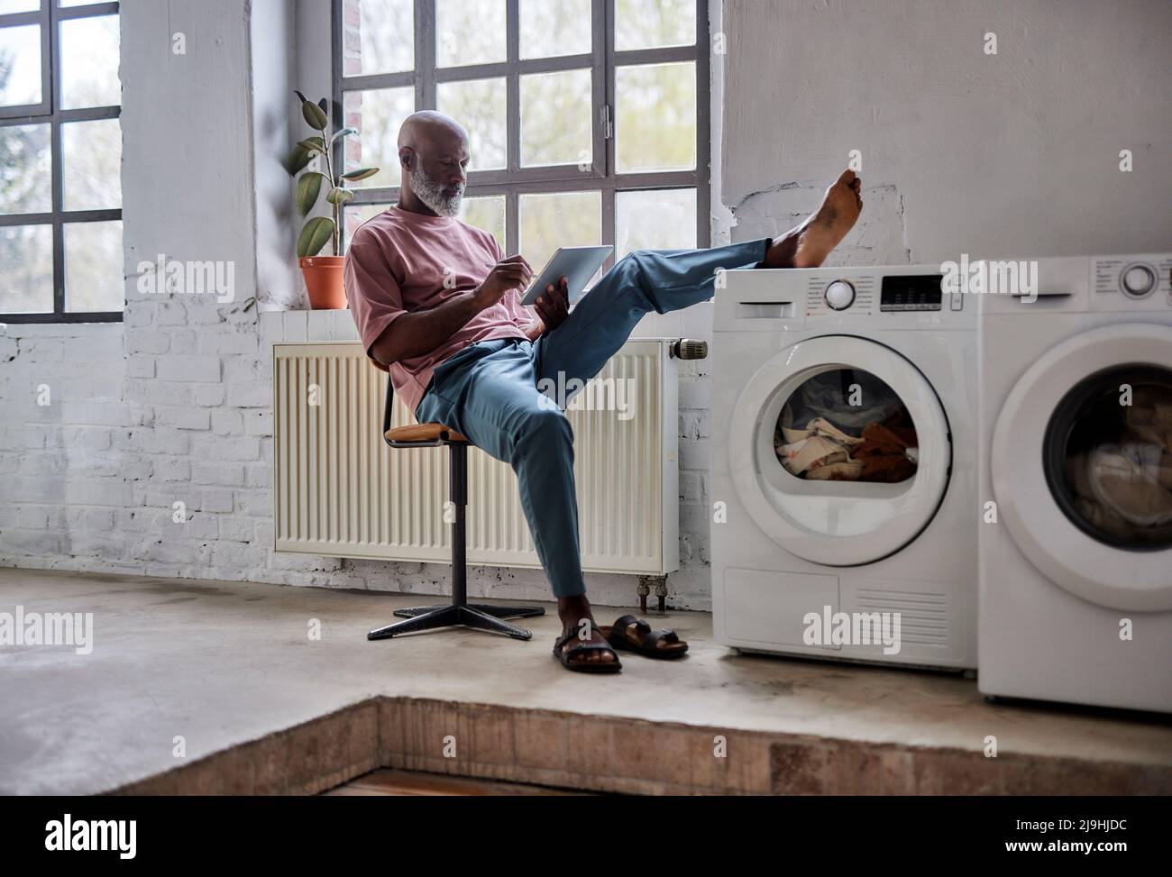 Mature man using tablet PC with foot on washing machine sitting on chair at home Stock Photo