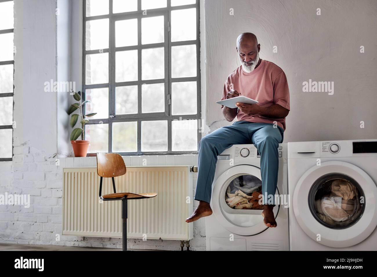 Bald man using tablet PC sitting on washing machine at home Stock Photo