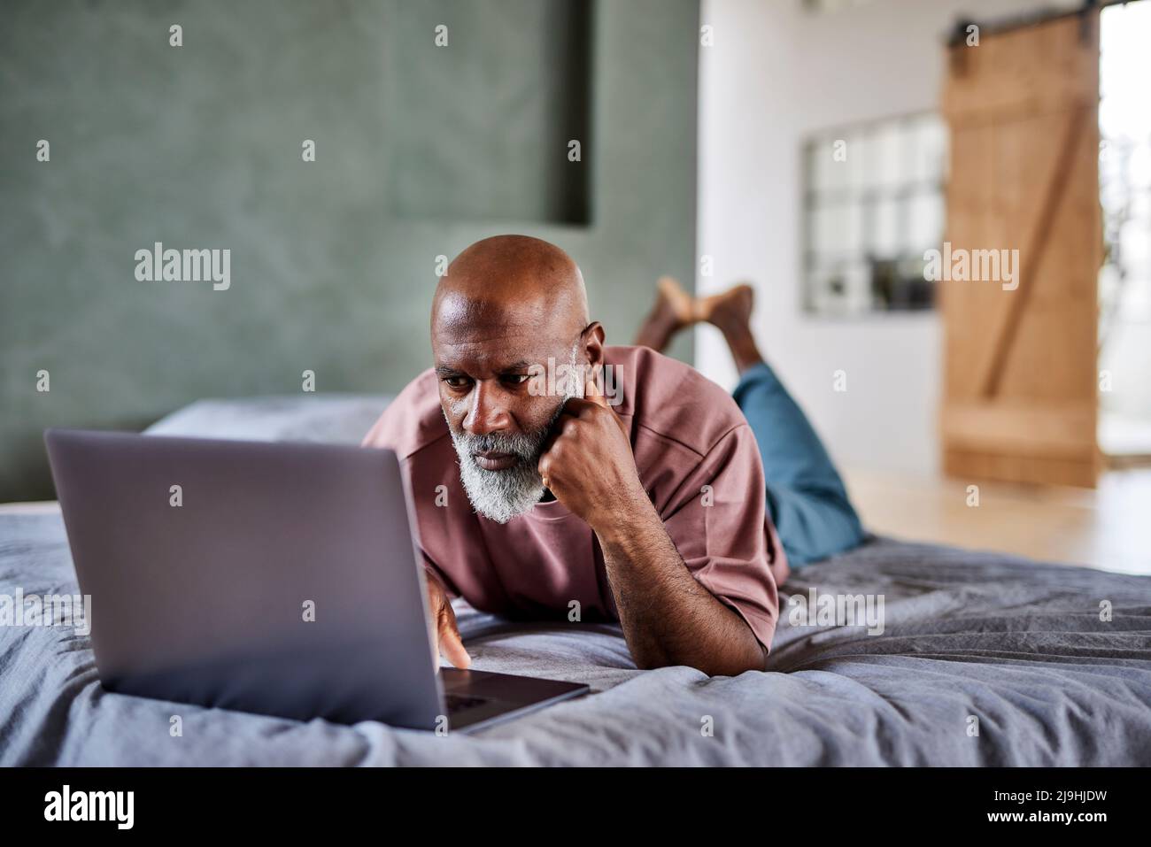 Mature man with white beard using laptop lying on bed at home Stock Photo