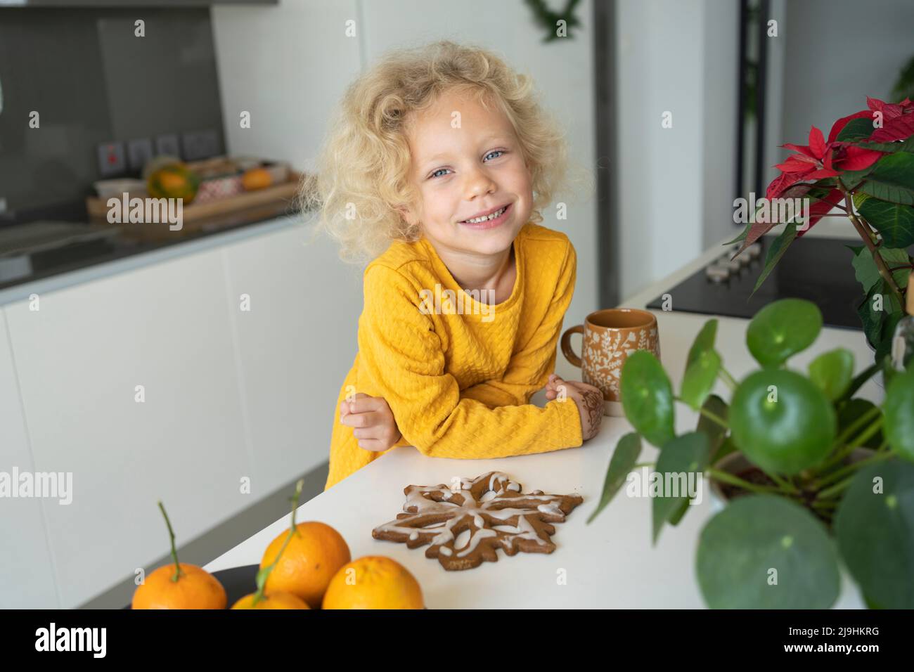 Smiling blond girl with gingerbread leaning on kitchen island Stock Photo