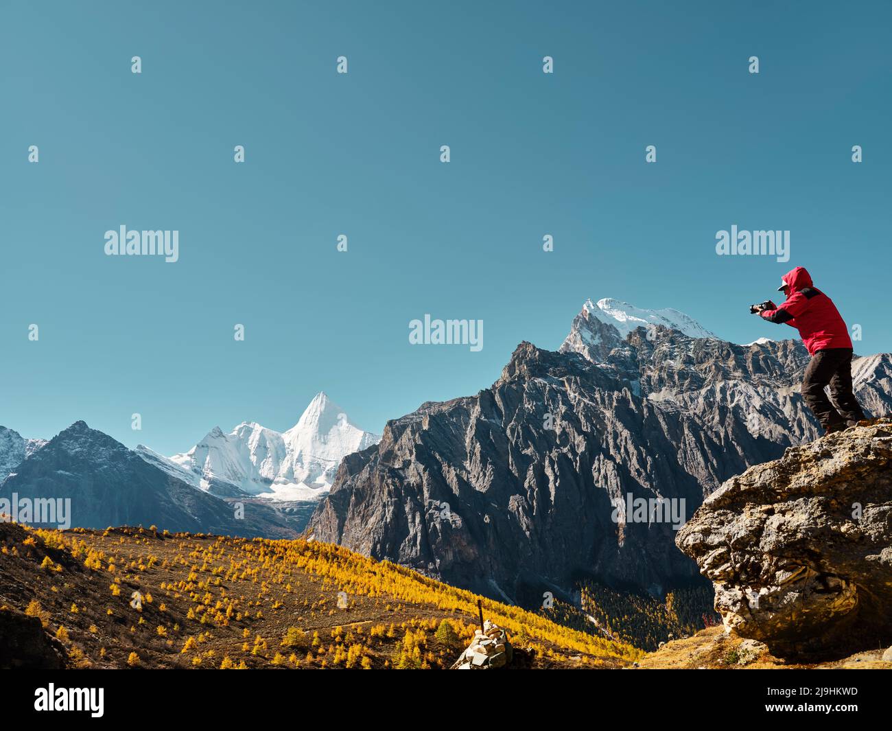 asian man standing on top of rock taking photo of Yangmaiyong (or Jampayang in Tibetan) mountain peak in the distance in Yading, Daocheng County, Sich Stock Photo