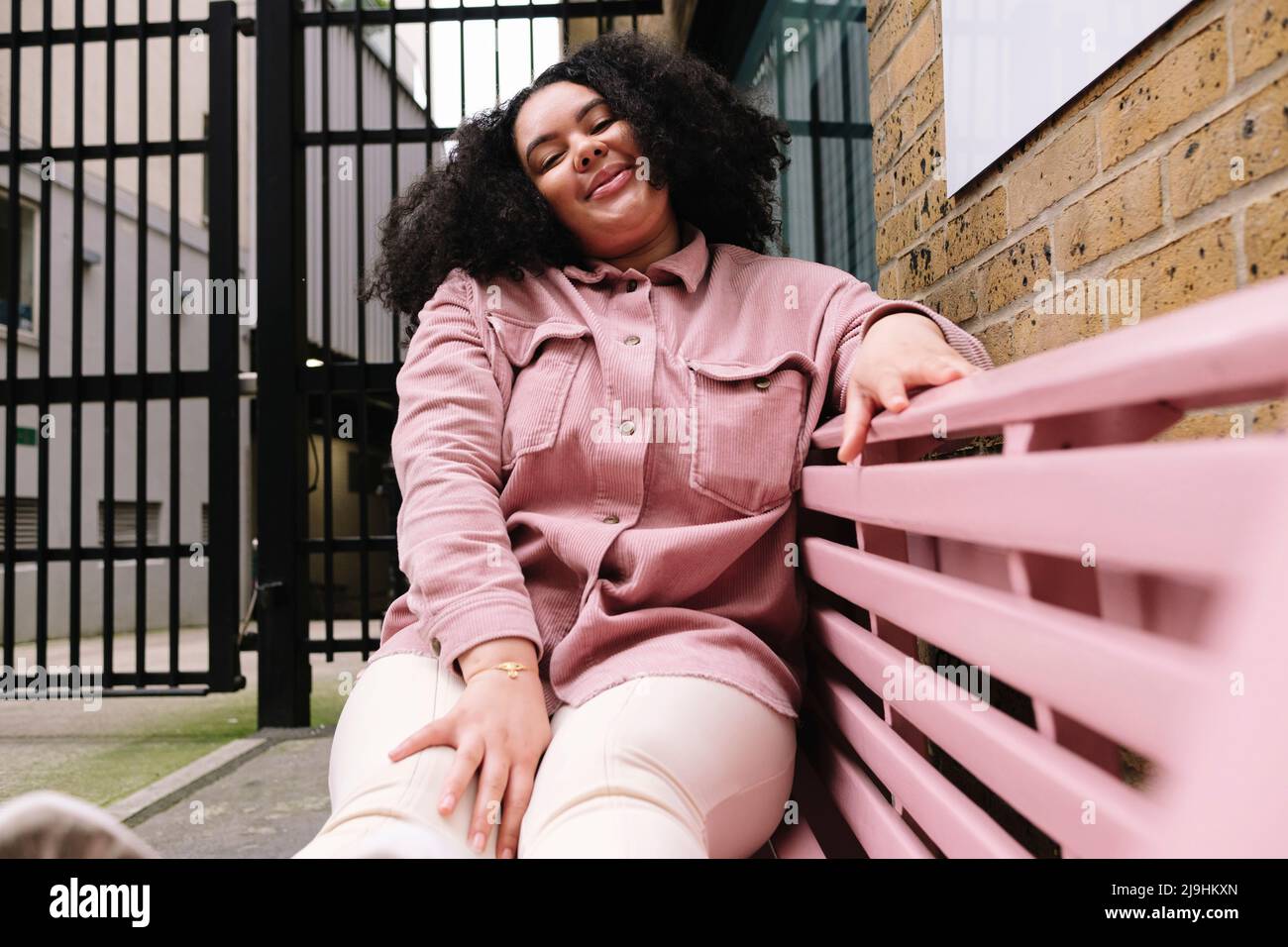 Smiling woman with curly hair sitting on pink bench Stock Photo