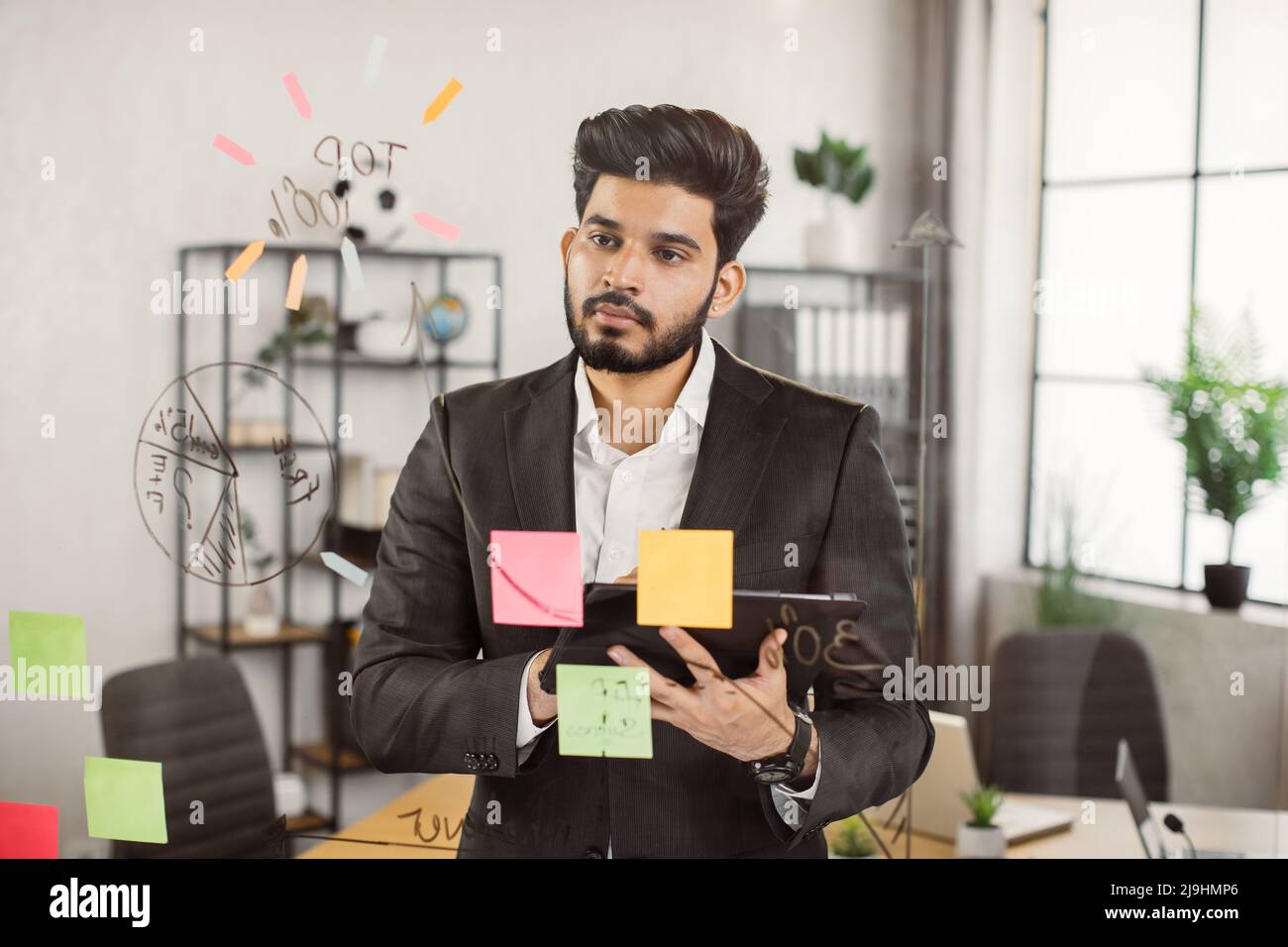 Corporation employee standing at modern office and looking at financial diagramm on glass board. Indian male worker in formal suit figuring out investment strategies with digital tablet in hands. Stock Photo