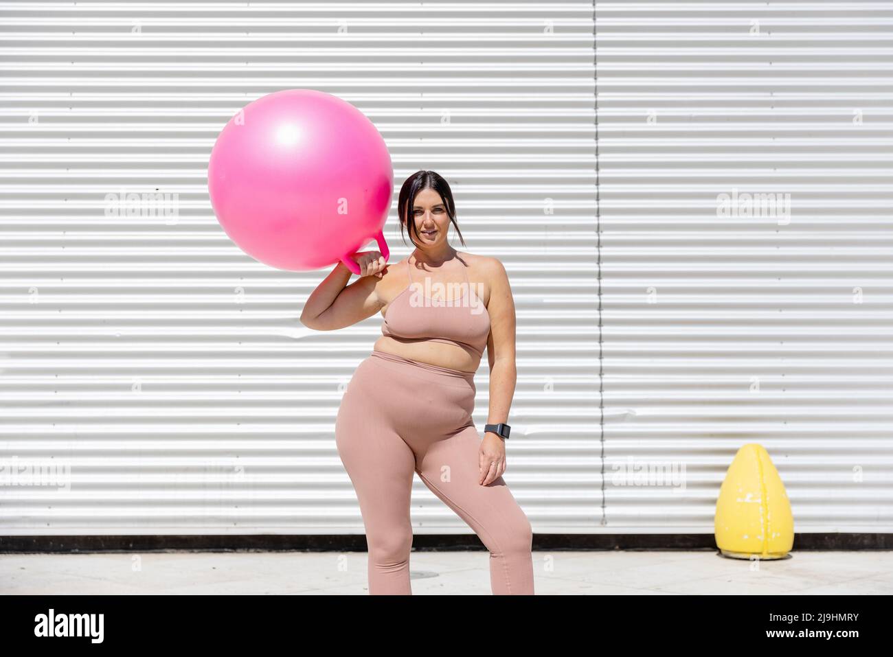 Curvy beautiful woman holding pink fitness ball standing in front of corrugated wall Stock Photo
