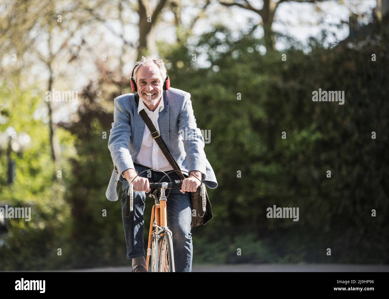 Smiling businessman listening music through wireless headphones cycling on sunny day Stock Photo