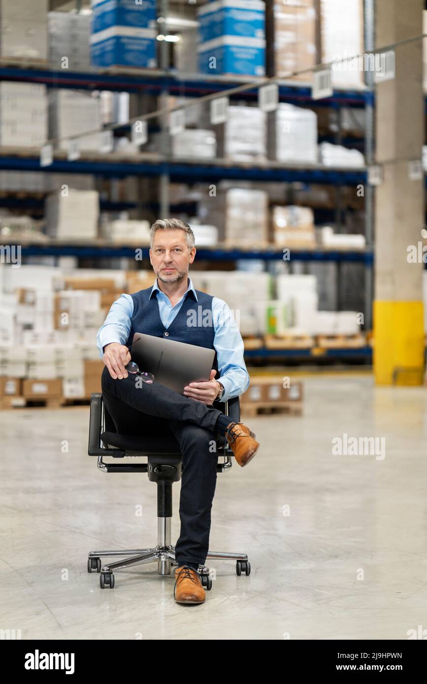 Confident mature businessman with legs crossed at knee and laptop in factory Stock Photo