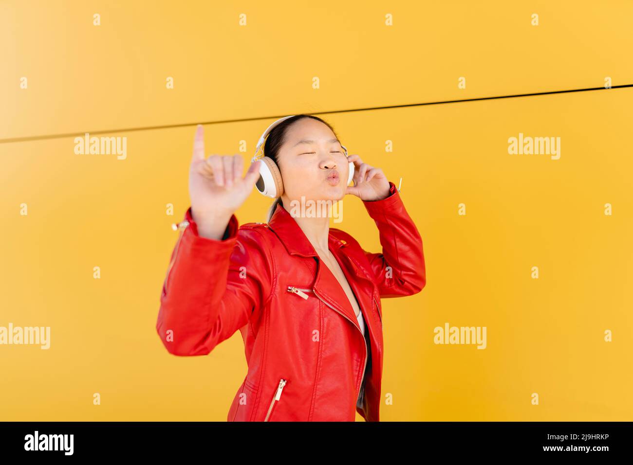 Young woman listening music through wireless headphones showing Shaka sign in front of yellow wall Stock Photo