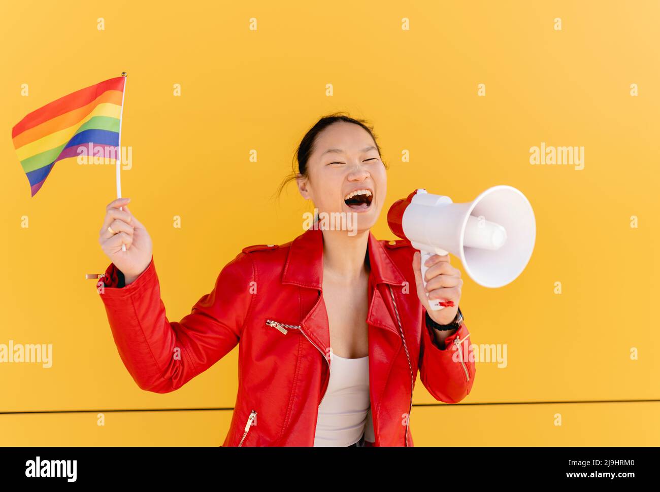 Happy woman holding rainbow flag shouting through megaphone in front of yellow wall Stock Photo