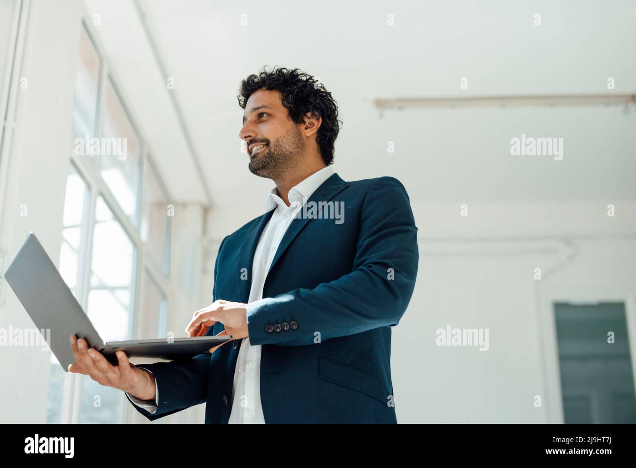 Smiling businessman holding laptop standing in office Stock Photo