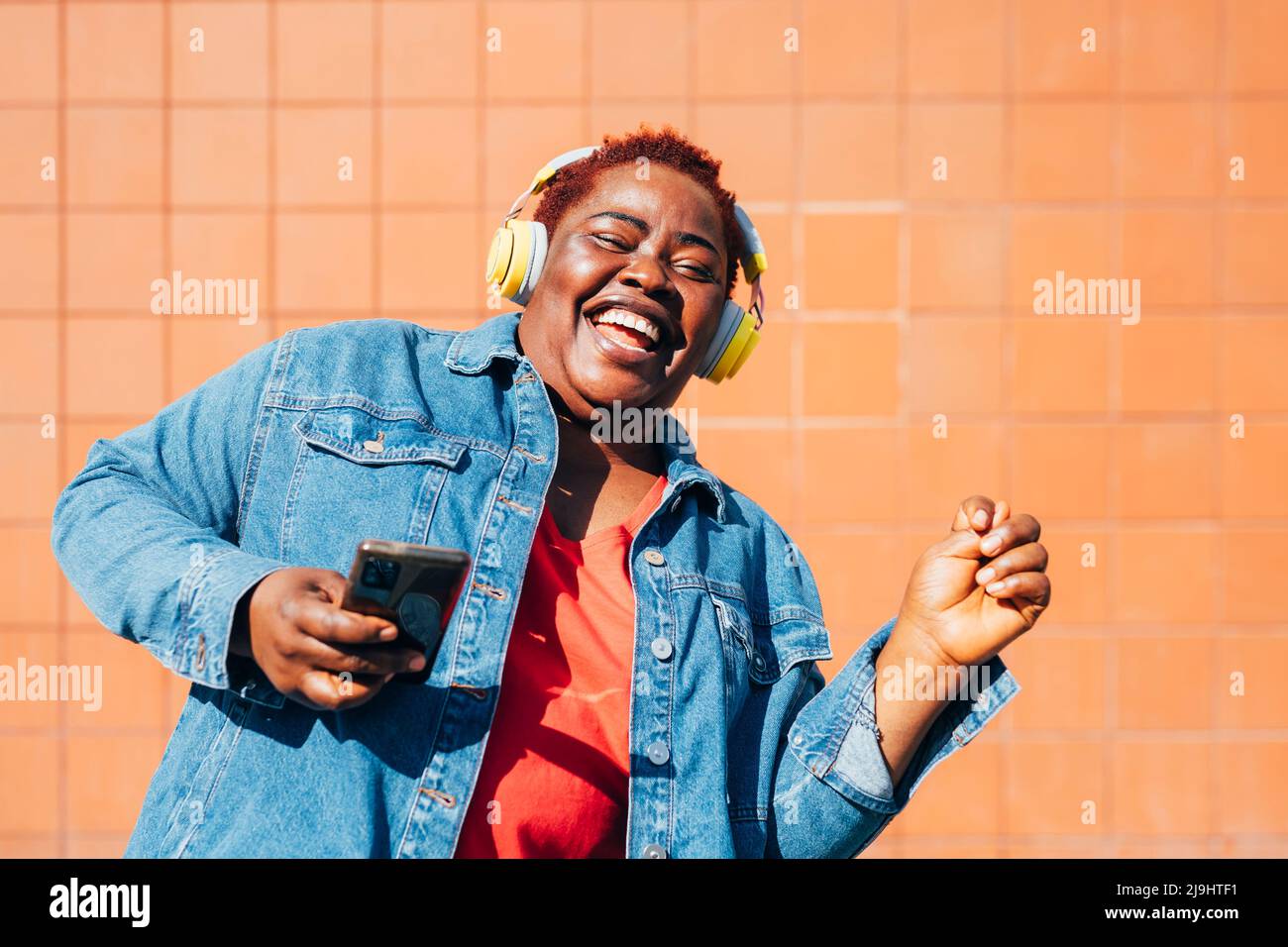 Cheerful woman holding smart phone listening music through wireless headphones enjoying in front of wall Stock Photo