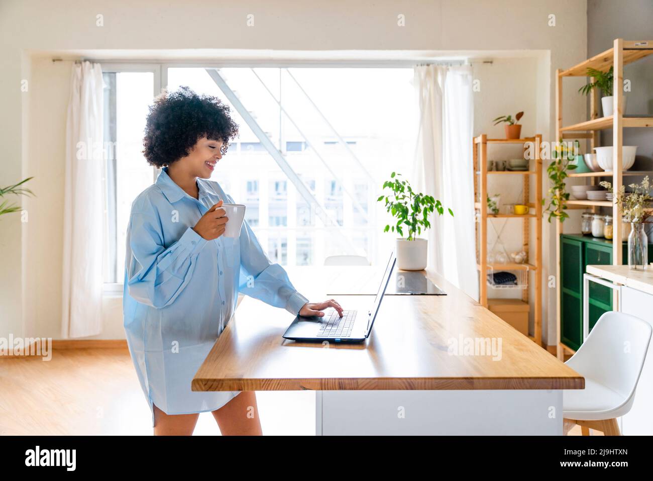 Happy woman with coffee cup using laptop at kitchen island Stock Photo