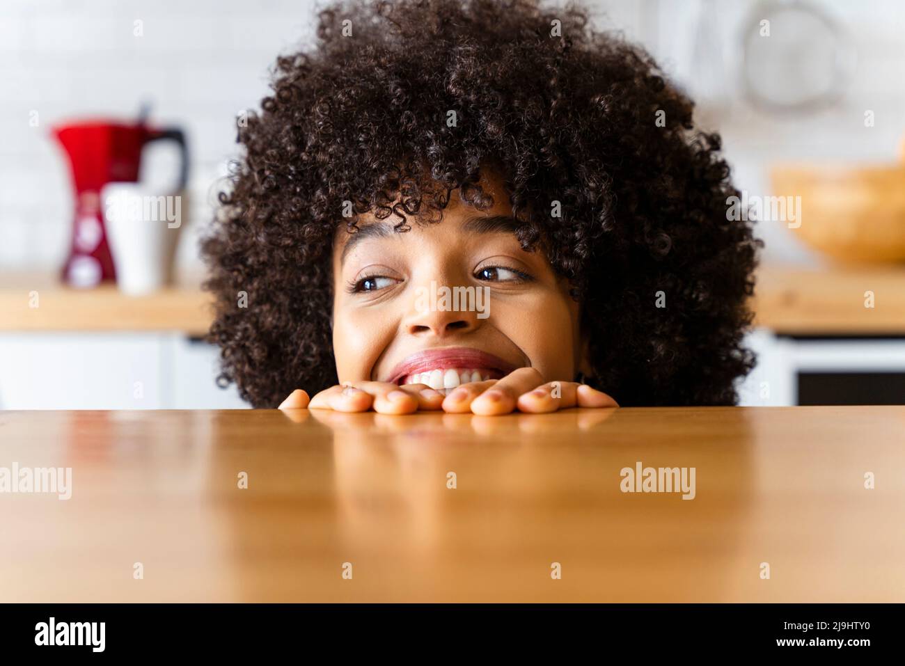Happy woman with Afro hairstyle hiding behind kitchen island at home Stock Photo
