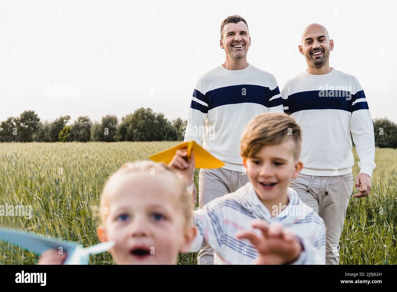 Gay male couple having fun with sons children outdoor - LGBTQ family playing with airplane paper toy - Focus on fathers face Stock Photo