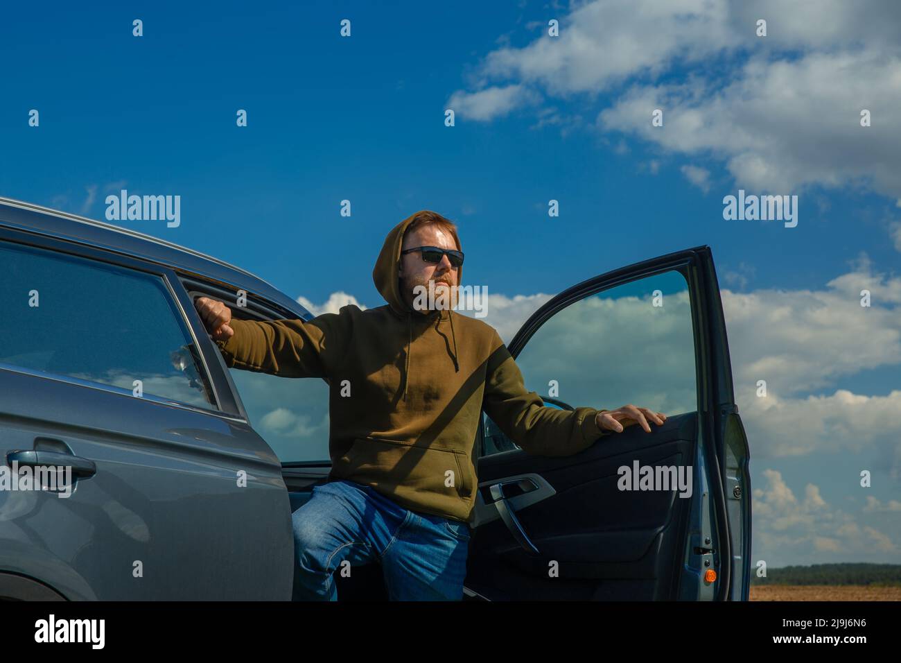 Young man weared in hoody and sun glasses holding his luxury car door and looking far away. Stock Photo