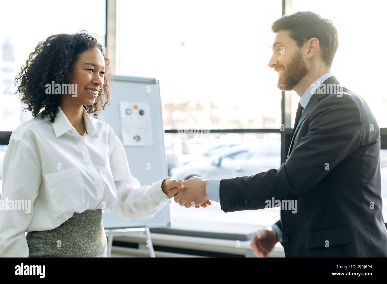 Successful influential business colleagues, young adult Caucasian man and African American woman, completed business negotiations, made a good deal, signed a contract, greet each other, shake hands Stock Photo