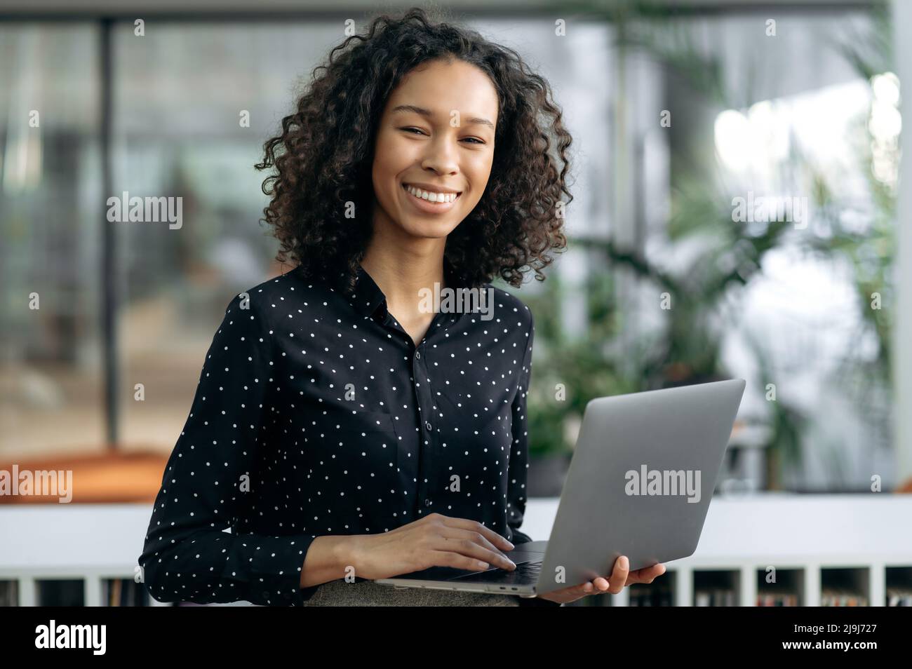 Portrait successful dark-skinned curly haired girl, financial analyst, auditor, IT specialist, stands in the office, in business clothes, holds an open laptop in her hands, looks at the camera, smiles Stock Photo