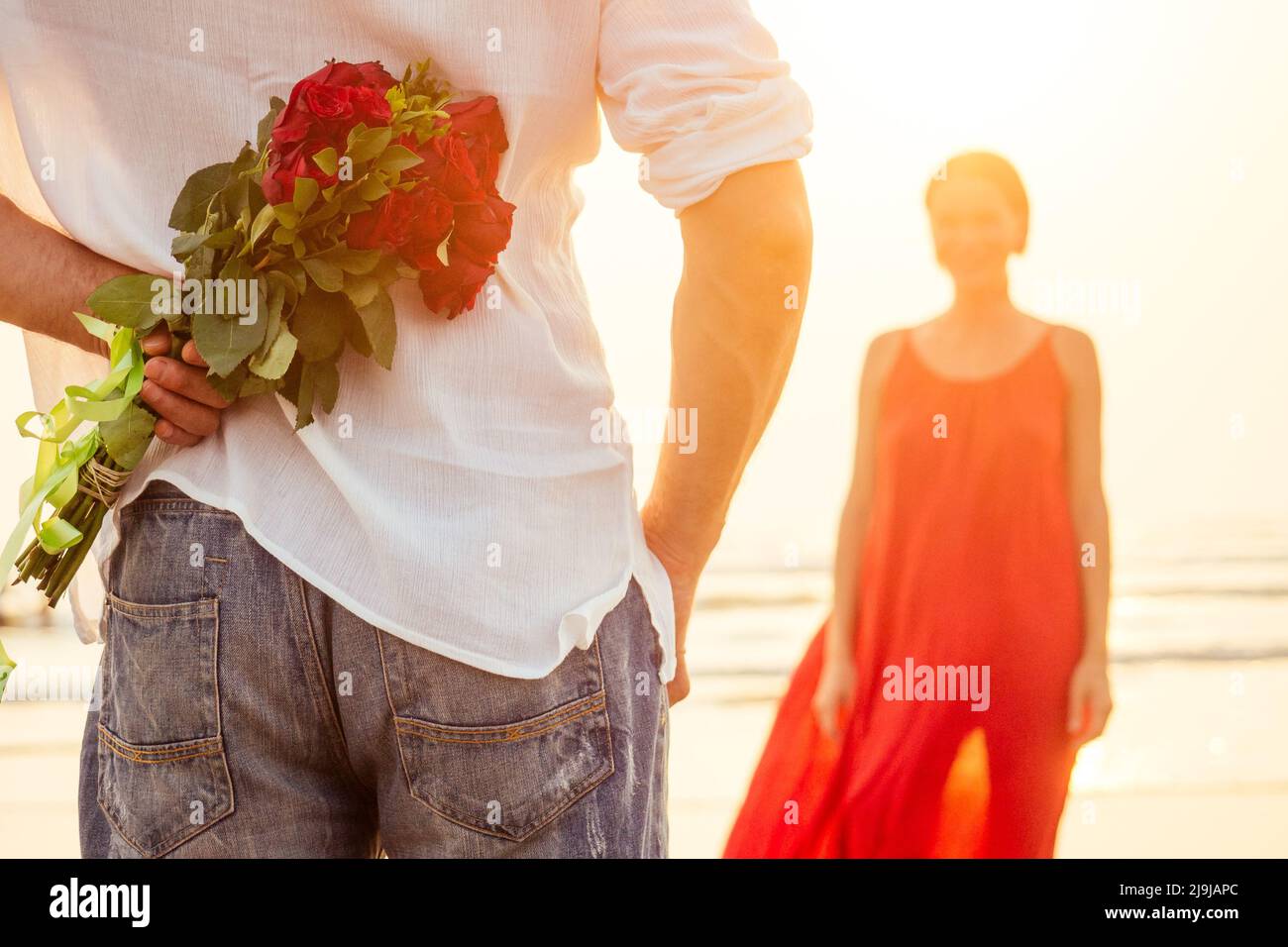 happy beautiful woman in red dress receiving a gift from a man by sea sunset.surprise wife from husband bouquet of roses women's day March 8 and Stock Photo