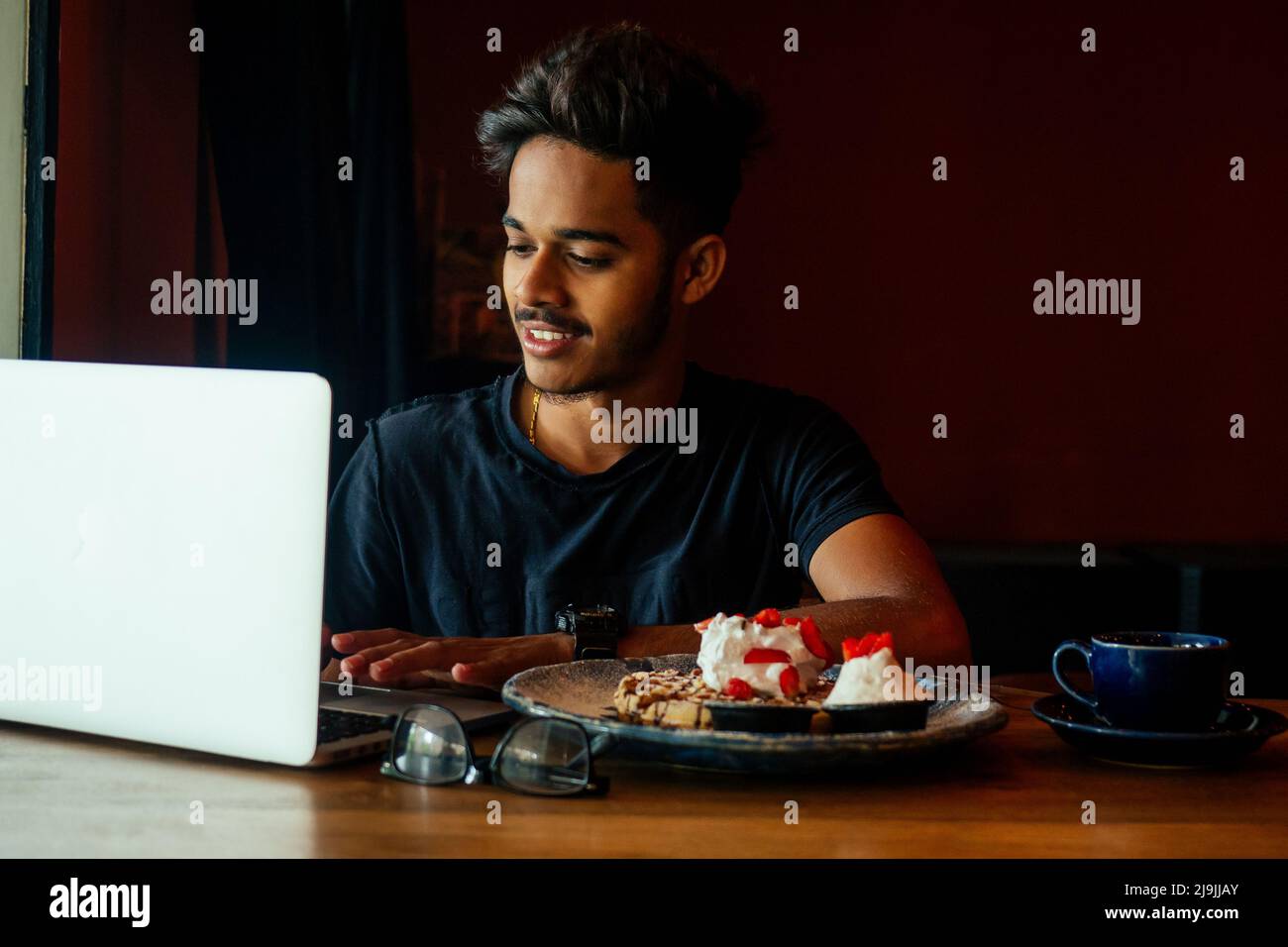Beard asian man in casual wear working with computer and eating cake with tea in cafe.student male chatting social network doing homework Stock Photo