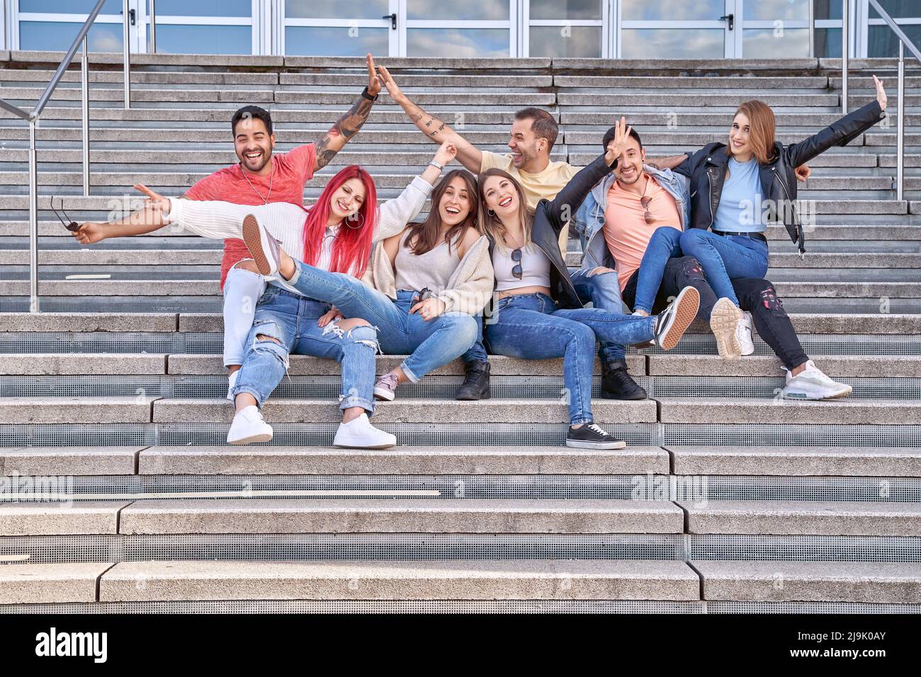 Group of cheerful multi-ethnic young men and women in casual outfits, smiling happily and looking at the camera as they sit on the stairs and hug each other after their successful studies. Stock Photo