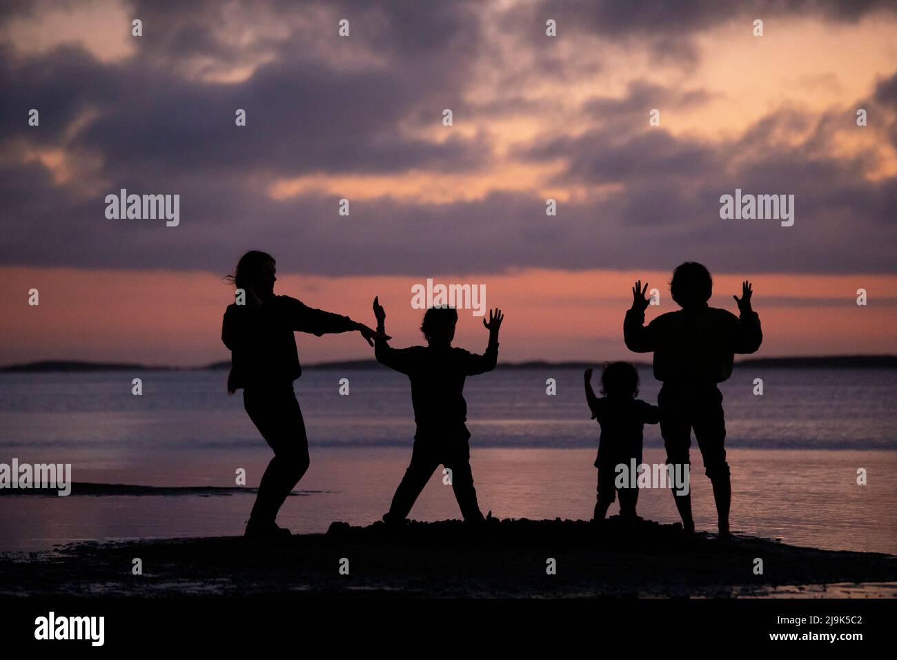Silhouetted family dancing, gesturing on ocean beach at sunset Stock Photo
